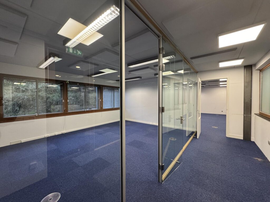 Modern empty office space on De Ruijterkade with blue carpet, glass partition walls, and large windows.